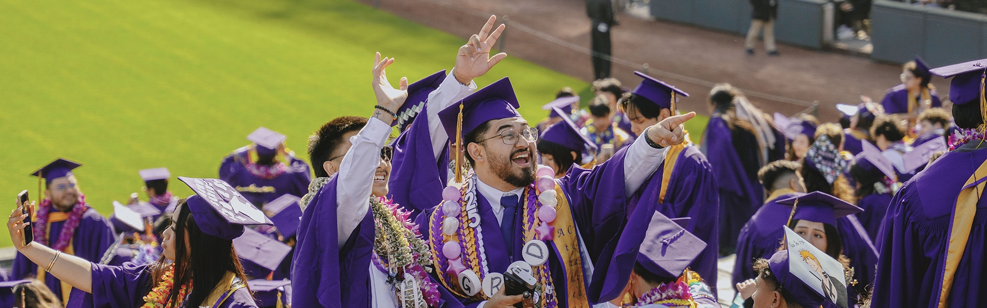 Two smiling students dressed in regalia inside Oracle Park stands stand and wave.